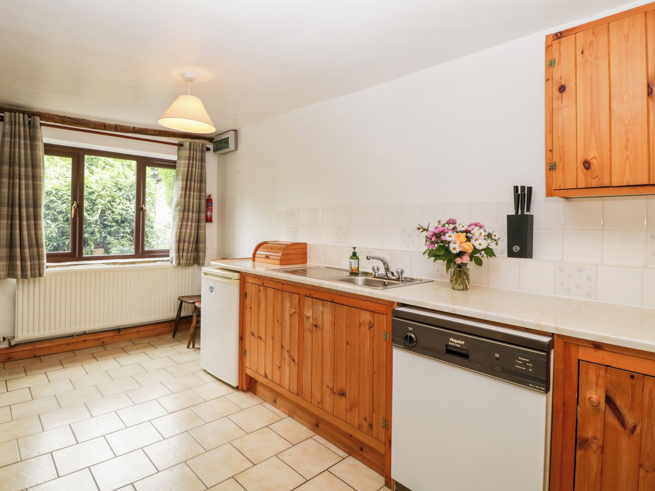 Une cuisine avec des meubles de rangement en bois, un plan de travail blanc, un lave-vaisselle encastré et une fenêtre avec des rideaux.