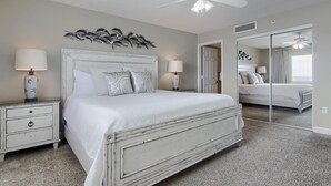 Une chambre à coucher avec un lit blanc, deux tables de chevet avec des lampes, un ventilateur de plafond et un grand miroir.
