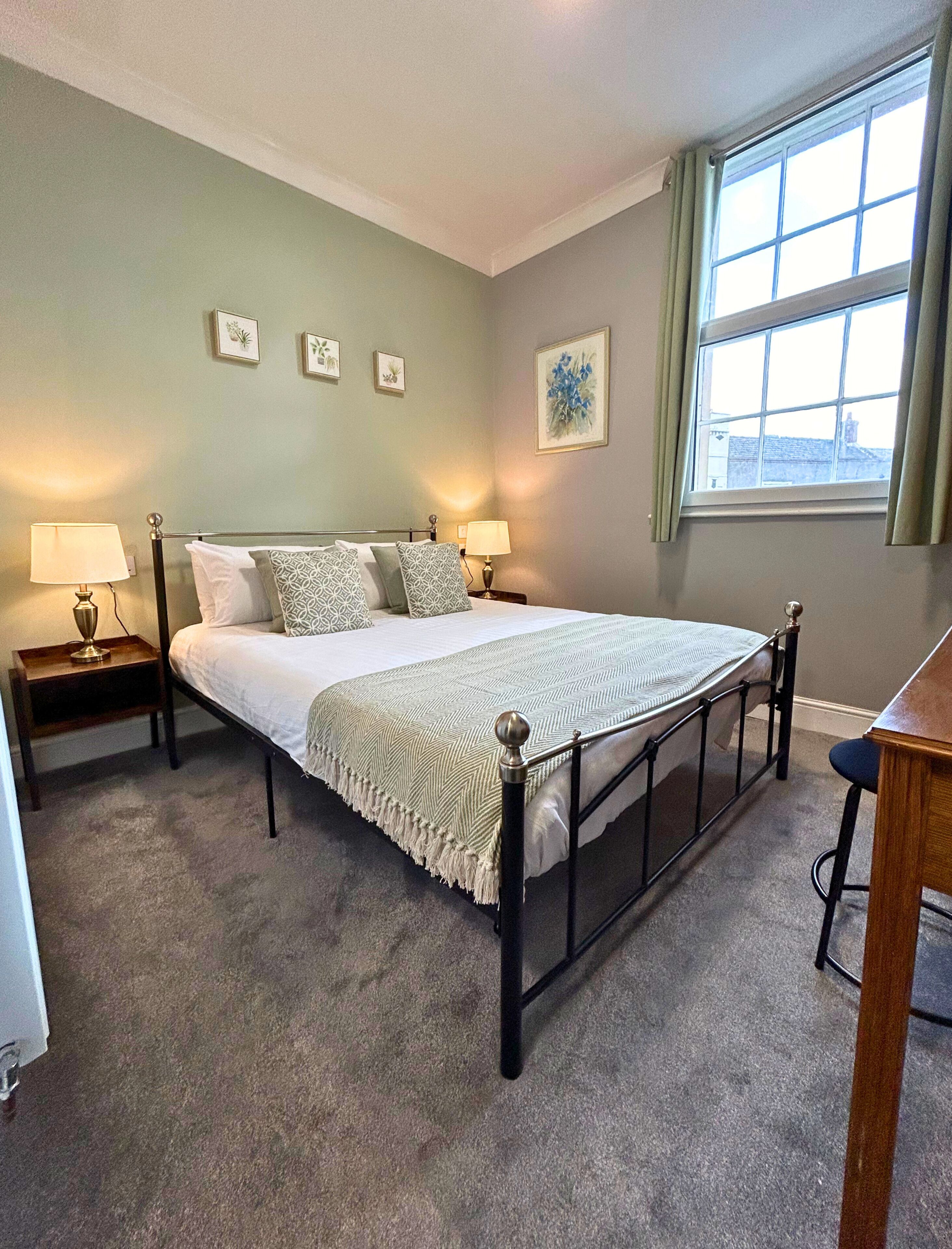A neatly arranged bedroom with a large bed, two bedside tables with lamps, a window with curtains, and a framed picture on the wall.
