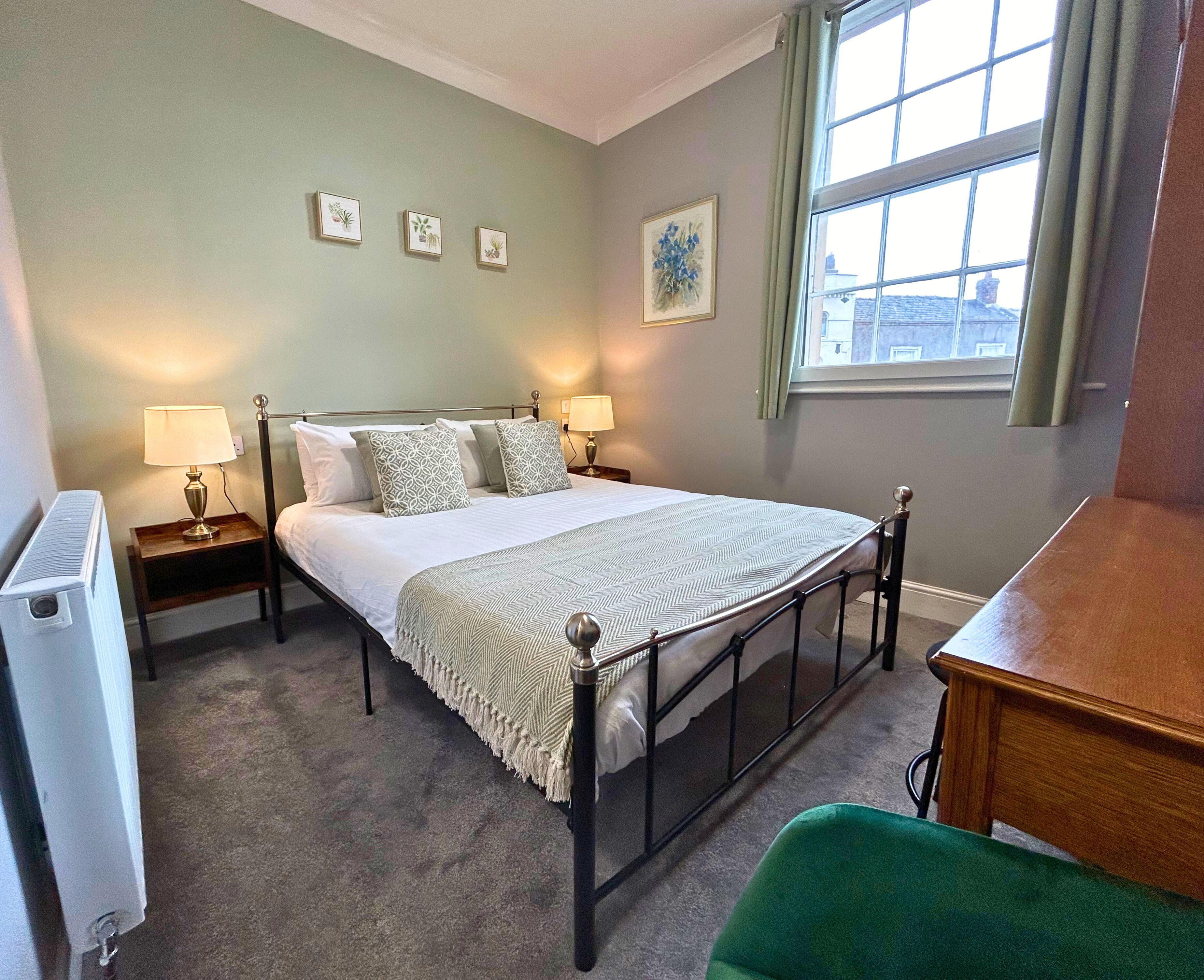 A neatly arranged bedroom with a bed, two bedside tables, a radiator, a window with curtains, and a green chair.