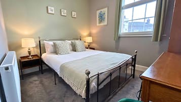 A neatly arranged bedroom with a bed, two bedside tables, a radiator, a window with curtains, and a green chair.