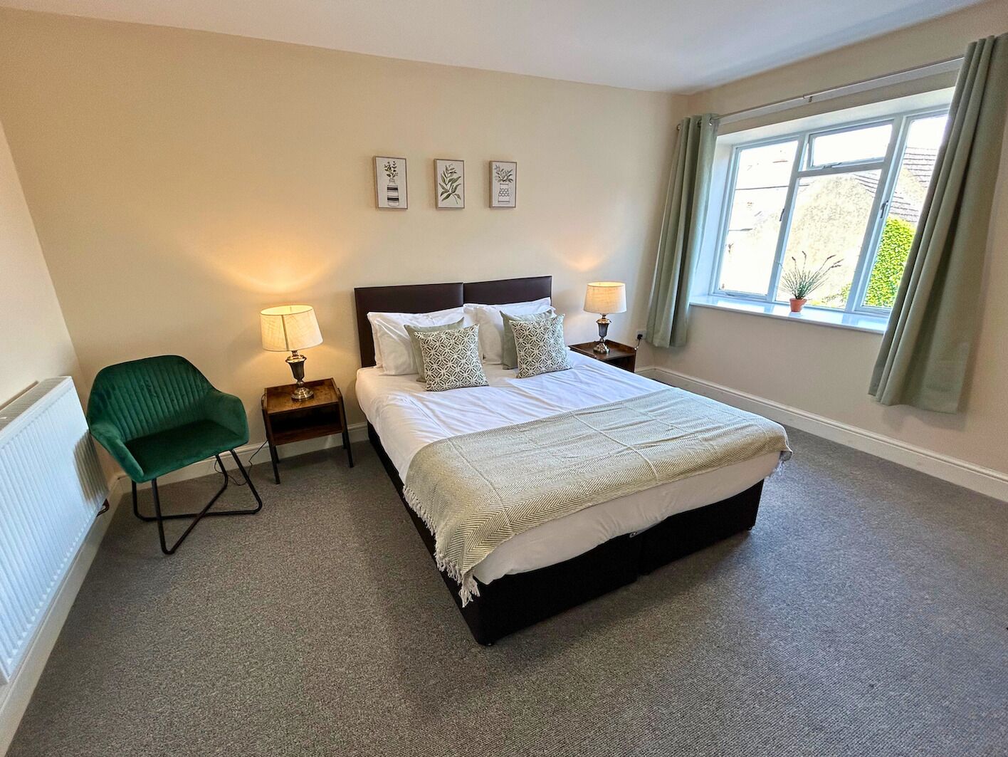 A bedroom with a bed, two bedside tables, a green chair, a window with curtains, and three wall-mounted pictures.