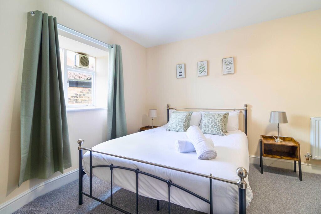 Une chambre à coucher bien rangée, avec un lit, deux tables de chevet, une fenêtre avec des rideaux et des œuvres d’art accrochées au mur.