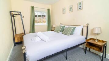 A neatly made bed with white linens and patterned pillows, a bedside table with a lamp, a wooden hanger stand, and a window with green curtains.