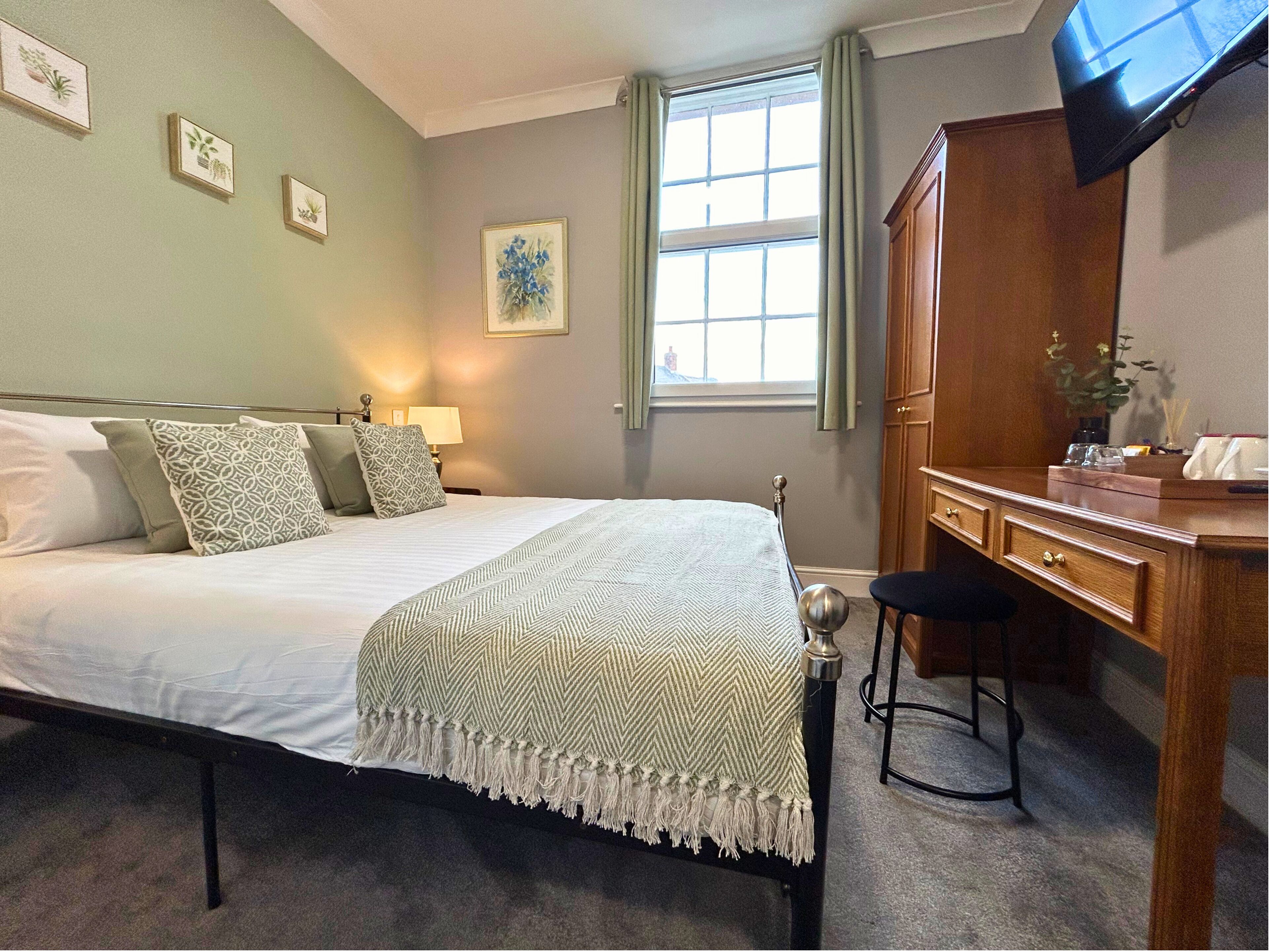 A neatly made bed with a patterned throw, a wooden desk with drawers, a television mounted on the wall, and a window with curtains.