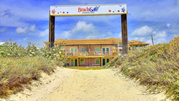 A beachfront hotel with a prominent BeachGate sign, multiple balconies, and a sandy pathway leading to the entrance. - Beachgate Condo Suites And Hotel 435 1 Bedroom Condo (Port Aransas)