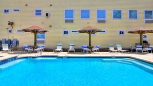 A swimming pool with blue water, surrounded by lounge chairs and umbrellas, against a yellow building facade with multiple windows. - Beachgate Condo Suites And Hotel 435 1 Bedroom Condo (Port Aransas)