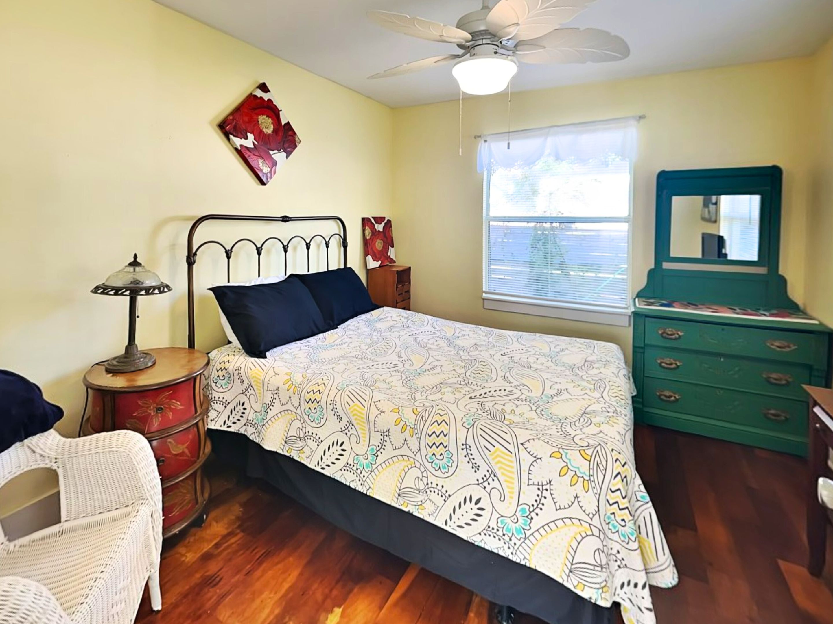 A bedroom with a bed, a nightstand, a dresser, a chair, a ceiling fan, and a window with blinds.
