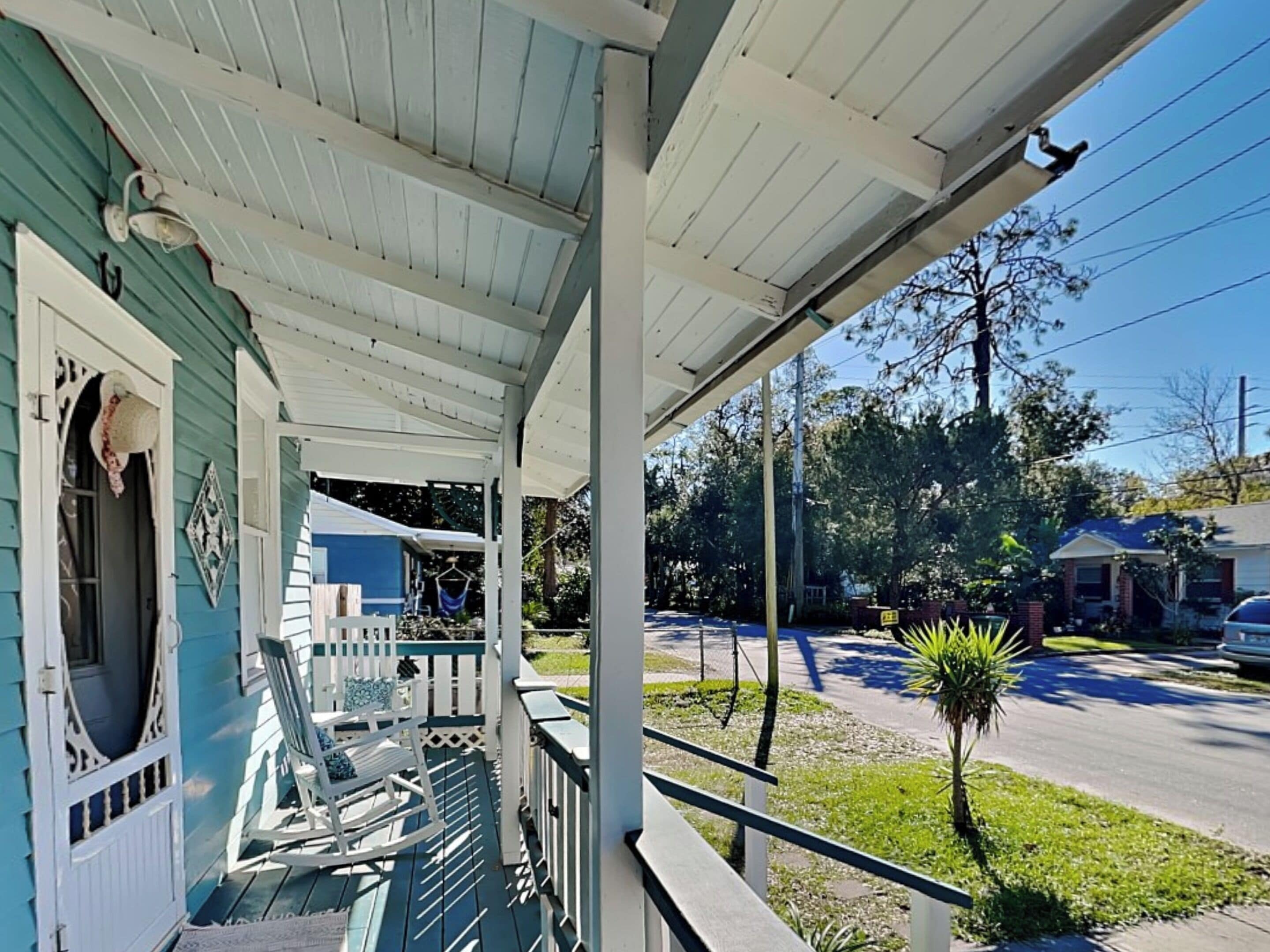 A porch with a rocking chair and a small table, overlooking a street with houses and a palm tree.