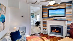 A living room with a fireplace, a television mounted on a wooden wall, a ceiling fan, and a view of the kitchen through an open door. - Cozy Casita by Distinctive Beach Rentals (St. Augustine)