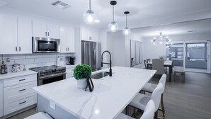Una cucina moderna con mobili bianchi, elettrodomestici in acciaio inossidabile, un'isola in marmo, illuminazione a sospensione e vista sull'area pranzo.