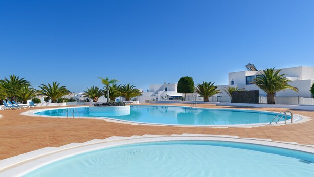 Une piscine avec une terrasse carrelée, des palmiers et des bâtiments modernes en arrière-plan.