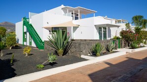 Un bâtiment moderne de couleur blanche, doté d’un escalier vert, entouré de plantes tropicales et surplombé d’un ciel bleu azur.
