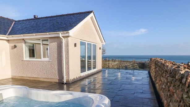 Ein modernes Haus mit Natursteinfassade, einem großen Panoramafenster und einem Whirlpool auf einer Terrasse mit Steinmauer und Meerblick.