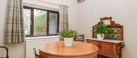 A dining room with a wooden table and chairs, a radiator, a window with curtains, and a wooden cabinet with a mirror and potted plants.