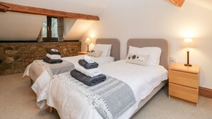 A cozy bedroom with two beds, stone walls, wooden beams, and a small wooden nightstand with a lamp. - Bullfinch Cottage (Leominster)