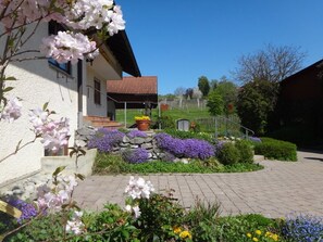 Un bâtiment à la façade en pierre, doté d’un toit recouvert de tuiles rouges et d’un jardin où poussent des fleurs violettes et blanches.