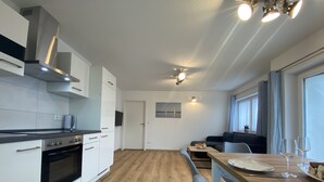 Eine moderne Küche mit weißen Schränken, einem eingebauten Ofen und einer Edelstahl-Dunstabzugshaube. Es gibt einen Essbereich mit einem Holztisch, Stühlen und einem kleinen Fenster.