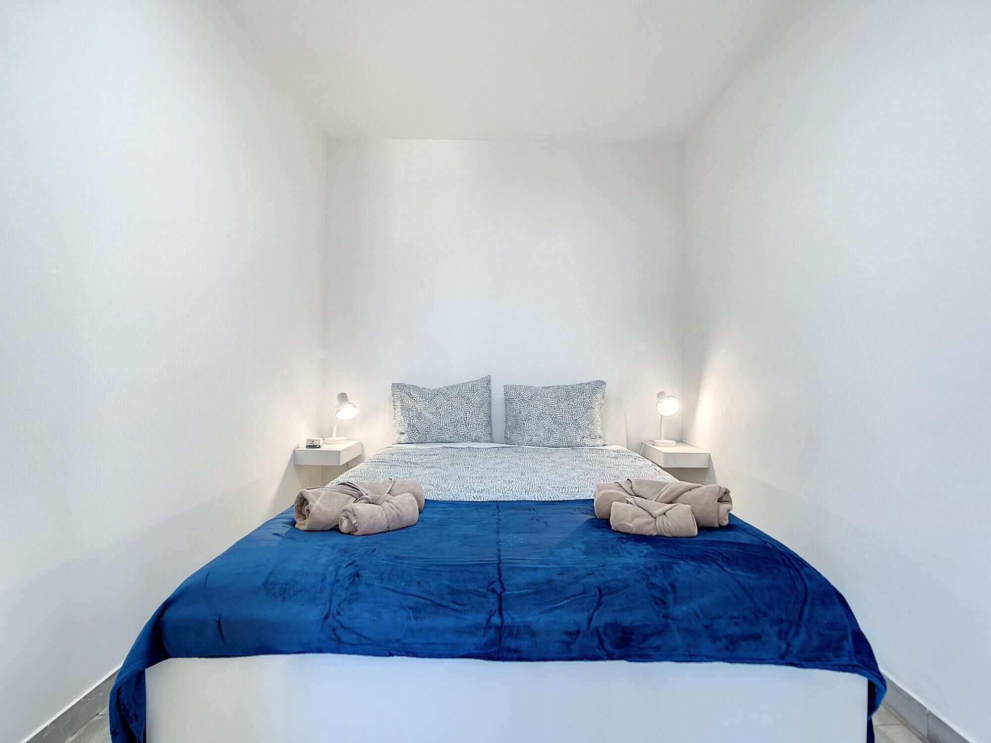A narrow, white-walled room with a bed at the center, flanked by two bedside tables with lamps.