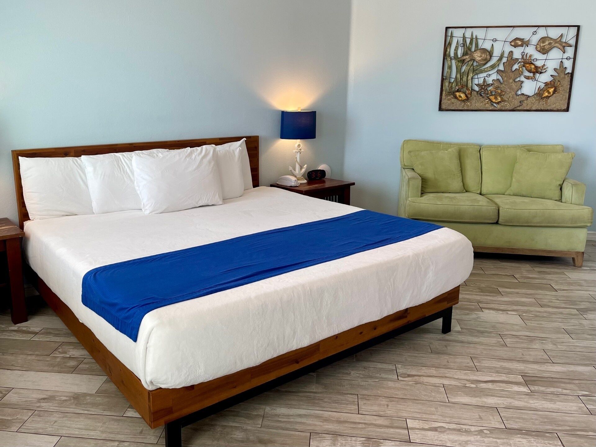 A neatly made bed with white and blue bedding, a bedside lamp, a clock, and a green sofa in a room with wooden flooring.