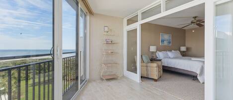 A balcony with a view of the ocean, a bedroom with a bed, a nightstand, and a chair.