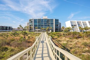 Un paseo marítimo que lleva a un edificio moderno con varios pisos y balcones, rodeado de palmeras y zonas verdes.