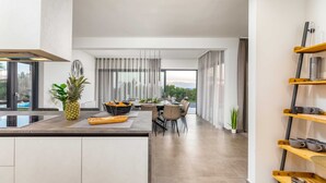 A modern kitchen with a central island, dining area with chairs, and a view of greenery outside. - Villa Borealis (Rasa)