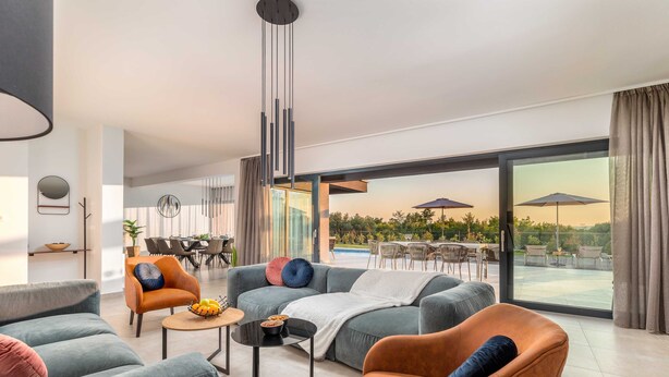 A modern living room with a large sofa, armchairs, a coffee table, and a dining area with a view of the outdoor patio. - Villa Borealis (Rasa)