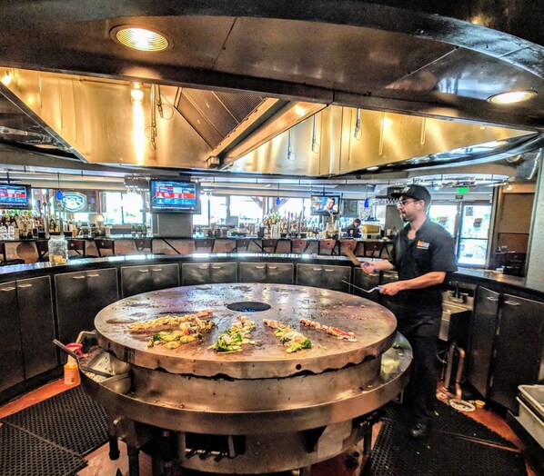 A chef preparing food on a large grill in a restaurant with multiple TVs and a bar area in the background.