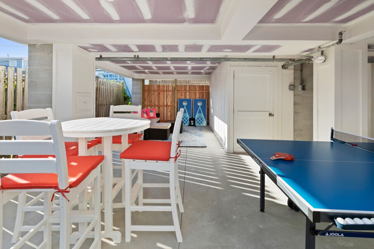A covered outdoor area with a ping pong table, white chairs with red cushions, and a view of a wooden fence and buildings in the background.