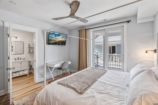 Un dormitorio con cama, escritorio, silla, ventilador de techo, ventana grande y balcón.