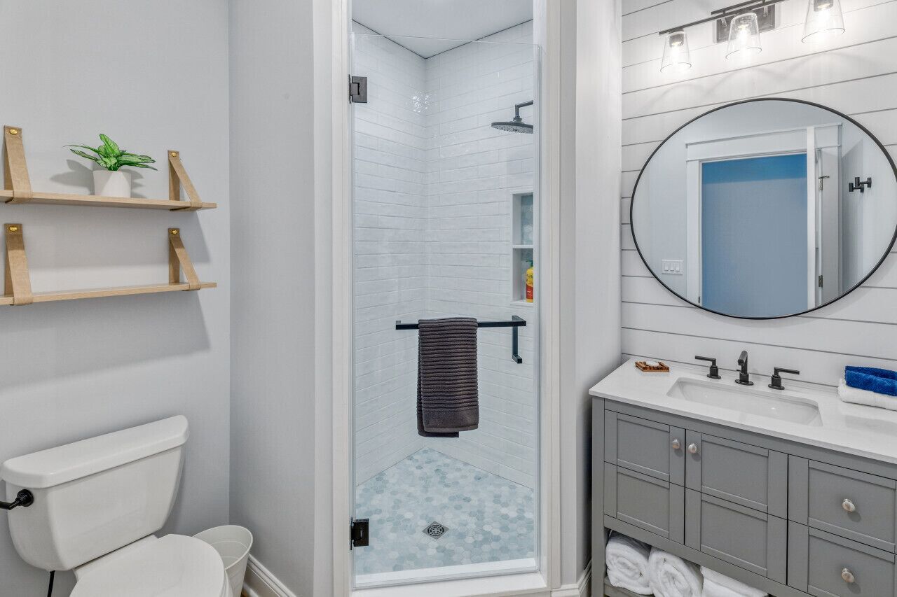 A bathroom with a toilet, a shower with a glass door, a sink with a mirror, and shelves with plants.