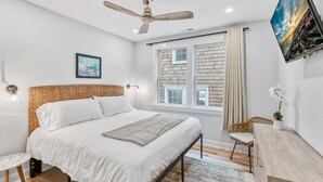 A bedroom with a large bed, a ceiling fan, a window with curtains, a desk with a chair, and a wall-mounted TV.