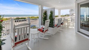 A covered outdoor area with white rocking chairs, a white fence, and a view of the beach.