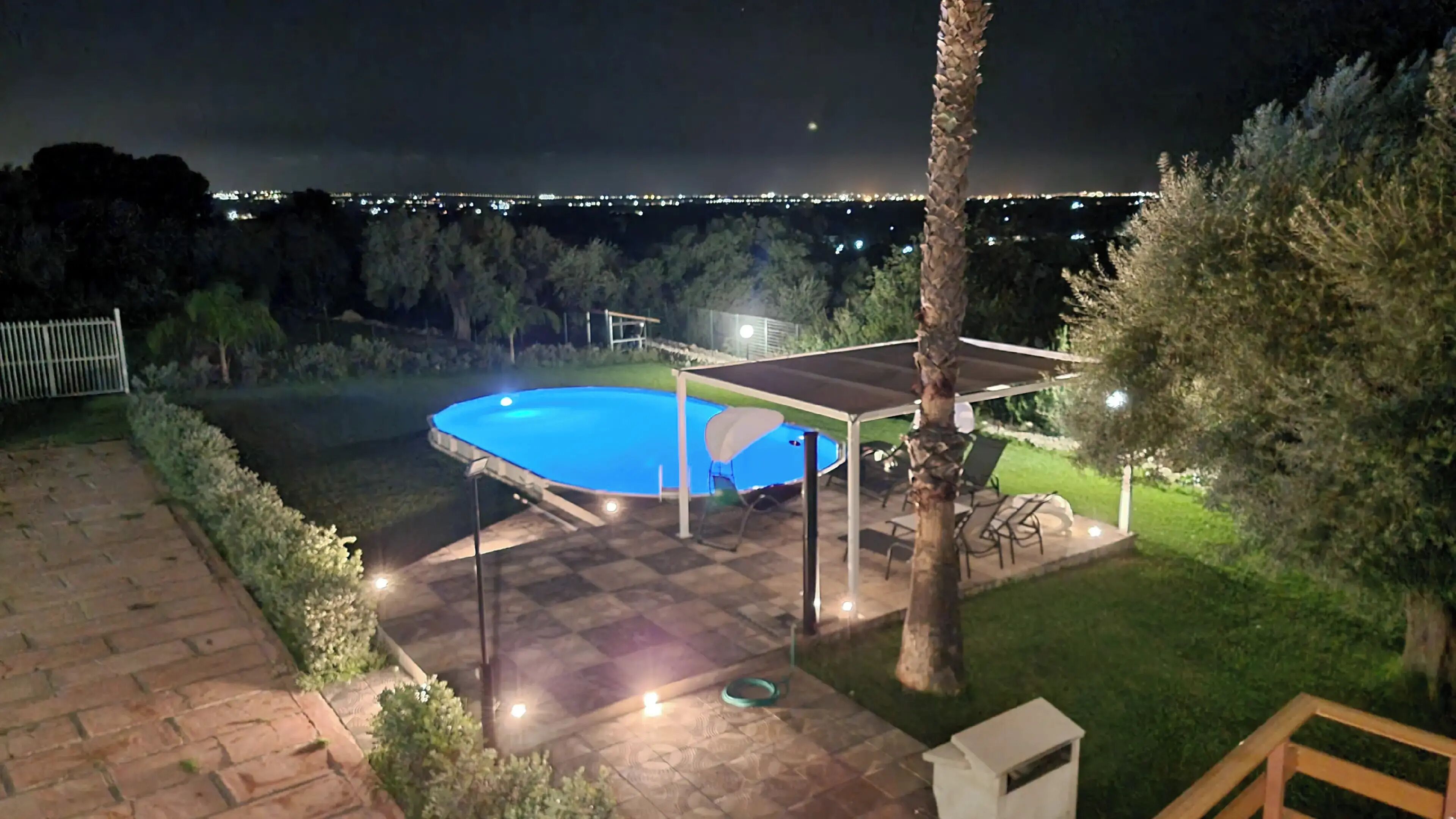A backyard with a swimming pool, a covered patio area, and a view of a cityscape at night.