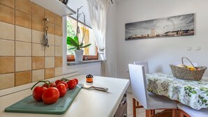 Una cocina con tabla de cortar, tomates, hervidor y ventana con cortinas.