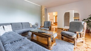 Una sala de estar con un sofá gris, una mesa de centro de madera y un sillón de madera. Hay una planta en maceta en la esquina.