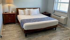 Un lit bien fait, recouvert d’une couverture à motifs, une lampe de chevet, une horloge numérique et une fenêtre avec des stores.