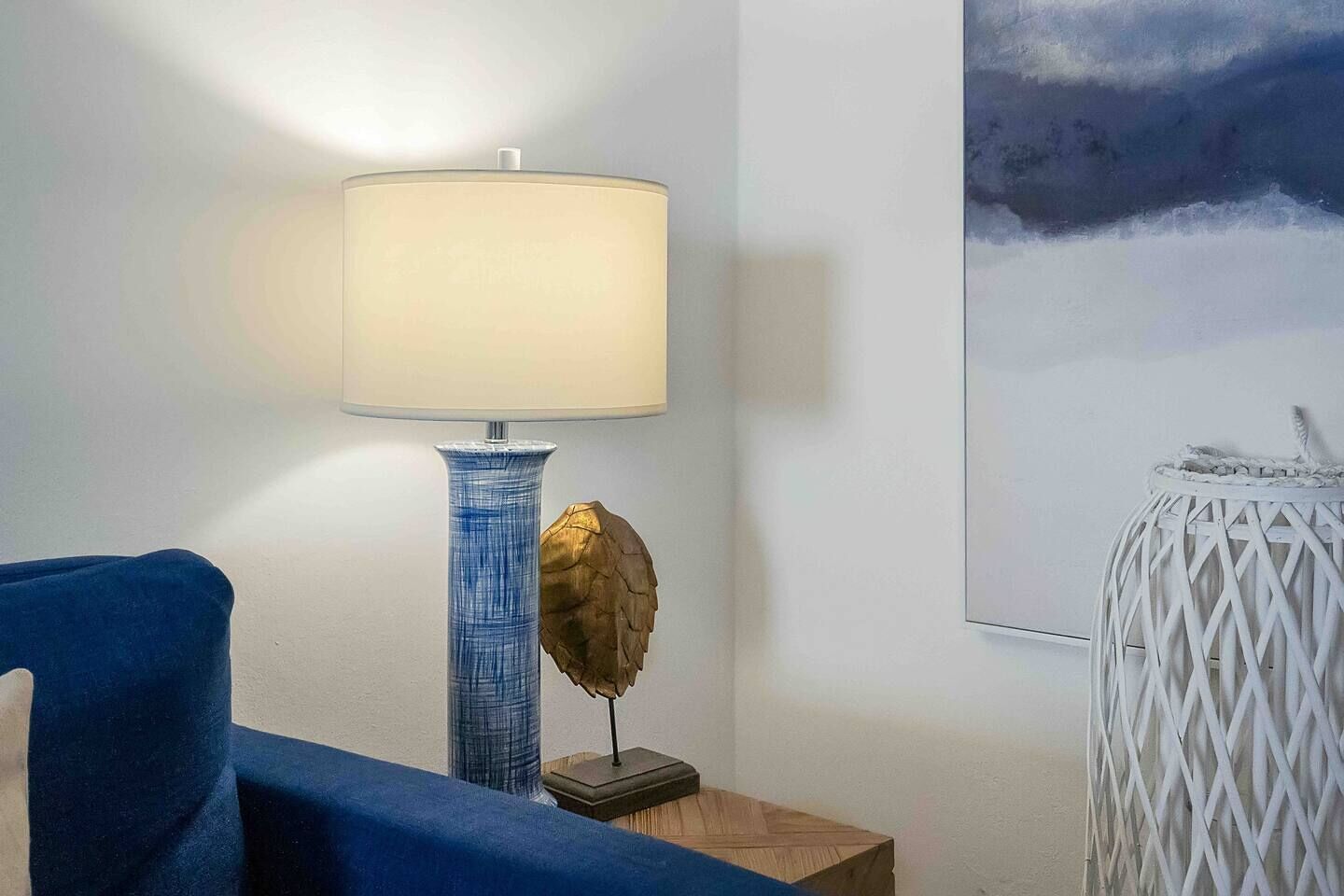 A room with a blue chair, a lamp with a blue vase, a small table with a golden leaf decoration, and a white woven lamp on a side table.