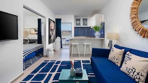 Un salon moderne avec un canapé bleu, une table basse et un téléviseur. Il y a également un coin cuisine avec un mur de crédence bleu et une vue sur l’extérieur.