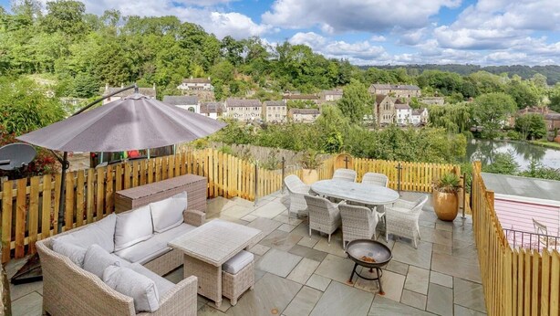A patio with wicker furniture, a table, and a grill, overlooking a village and a lake. - Dawns House in the Peaks overlooking the mill pond (Cromford)