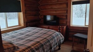 Ein Schlafzimmer in einer Blockhütte mit einem Bett, einem Fernseher auf einer Kommode und zwei Fenstern.
