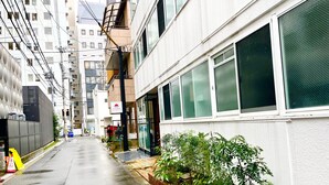 Una calle urbana estrecha flanqueada por edificios, con un letrero visible de un comercio y un pequeño jardín con plantas.