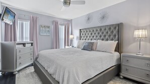 Une chambre à coucher avec un grand lit, un ventilateur de plafond, un téléviseur fixé au mur et une commode avec des tiroirs.