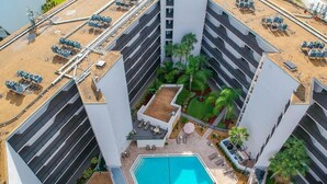 Una piscina en la azotea rodeada por un edificio moderno de varios pisos, con mobiliario exterior y un jardín ajardinado.