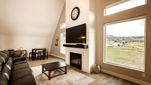 A living room with a fireplace, a television mounted above it, a large window offering a view of the countryside, and a leather sofa. - The spacious “LavaBarn” in Lava Hot Springs (Lava Hot Springs)