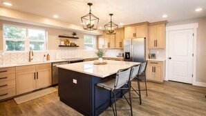 A modern kitchen with wooden cabinets, a central island with bar stools, pendant lighting, and a view of the outdoors through large windows. - The spacious “LavaBarn” in Lava Hot Springs (Lava Hot Springs)