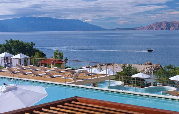 Relax by the pool overlooking the sea and distant mountains, with a small boat on the water and lush vegetation along the shoreline.