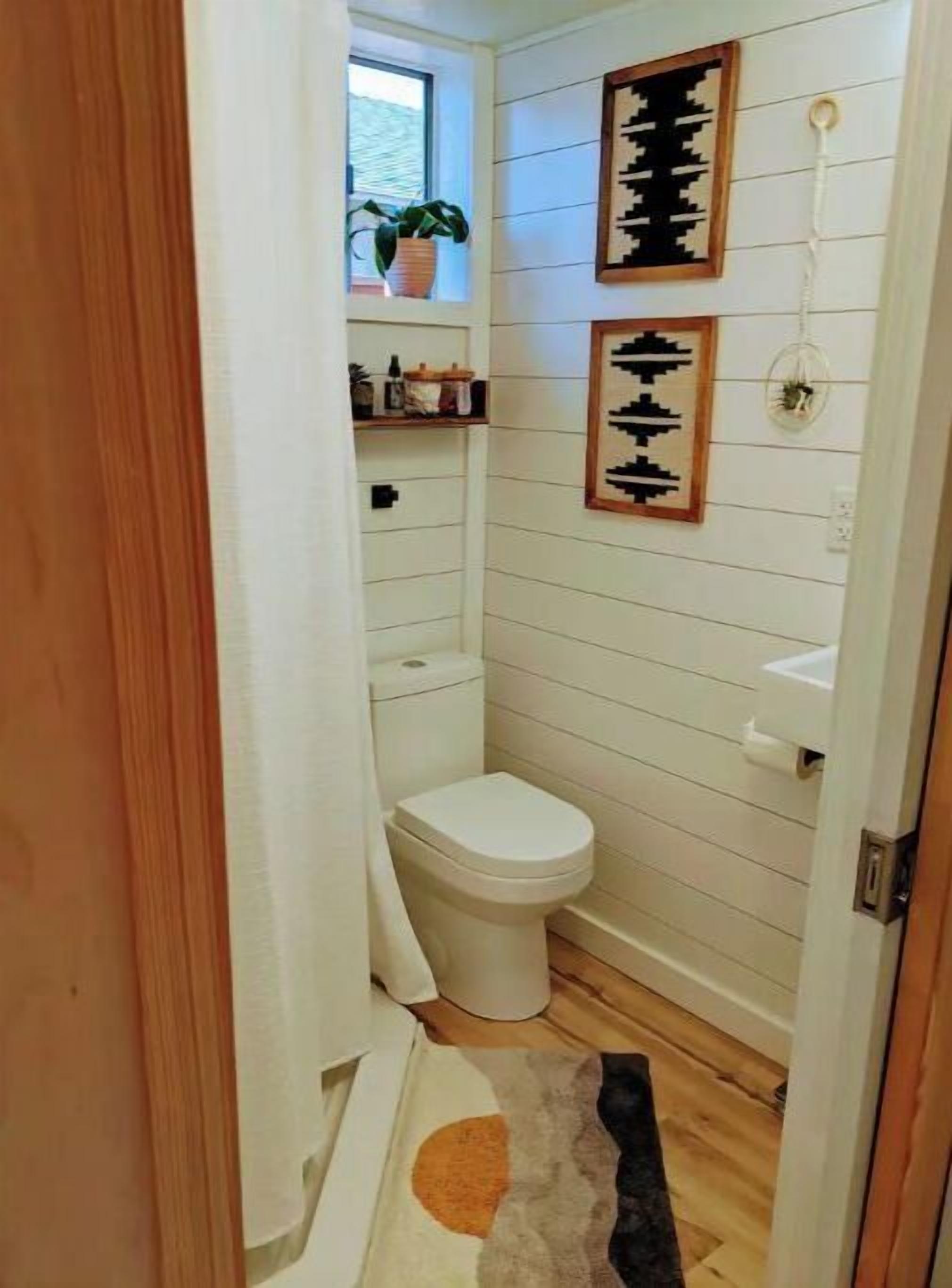 A narrow bathroom with a toilet, a shelf with toiletries, and a window with a plant.