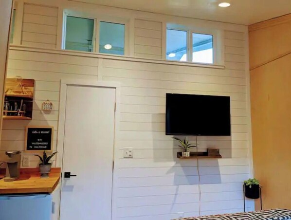 A room with a white paneled wall, a flat-screen TV mounted above a shelf, a door with a sign, and a small plant on the shelf.